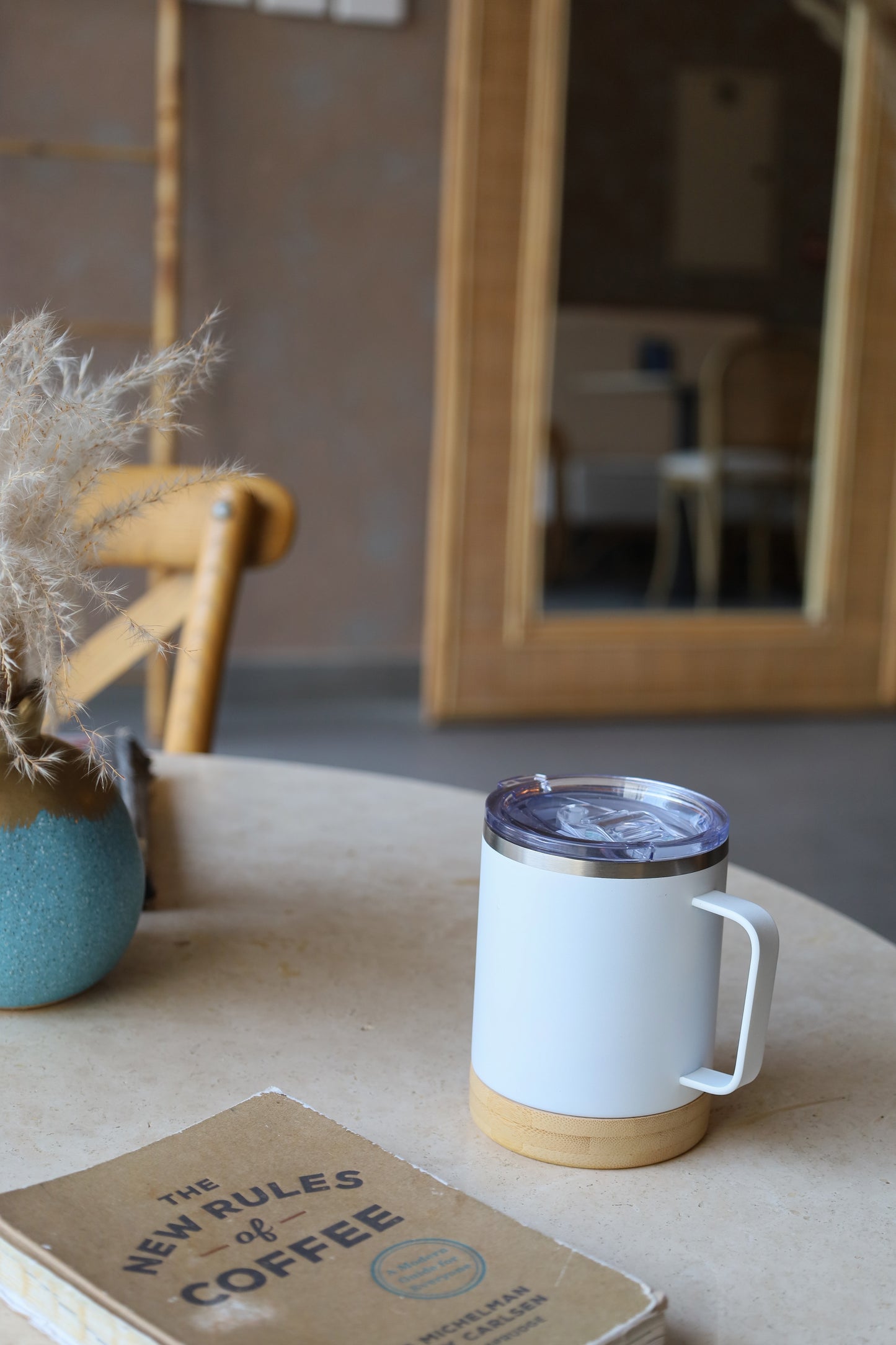 Bamboo Base Mug with white body and clear lid placed on wooden table next to a book and a blue vase with dried flowers