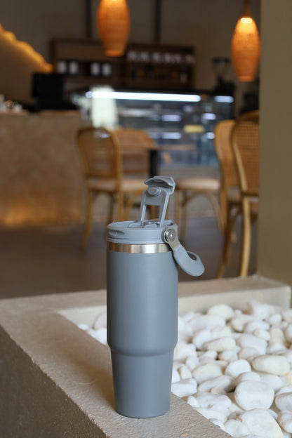Gray reusable Double Wall Stainless Steel Tumbler with flip lid placed on stone surface with white pebbles nearby