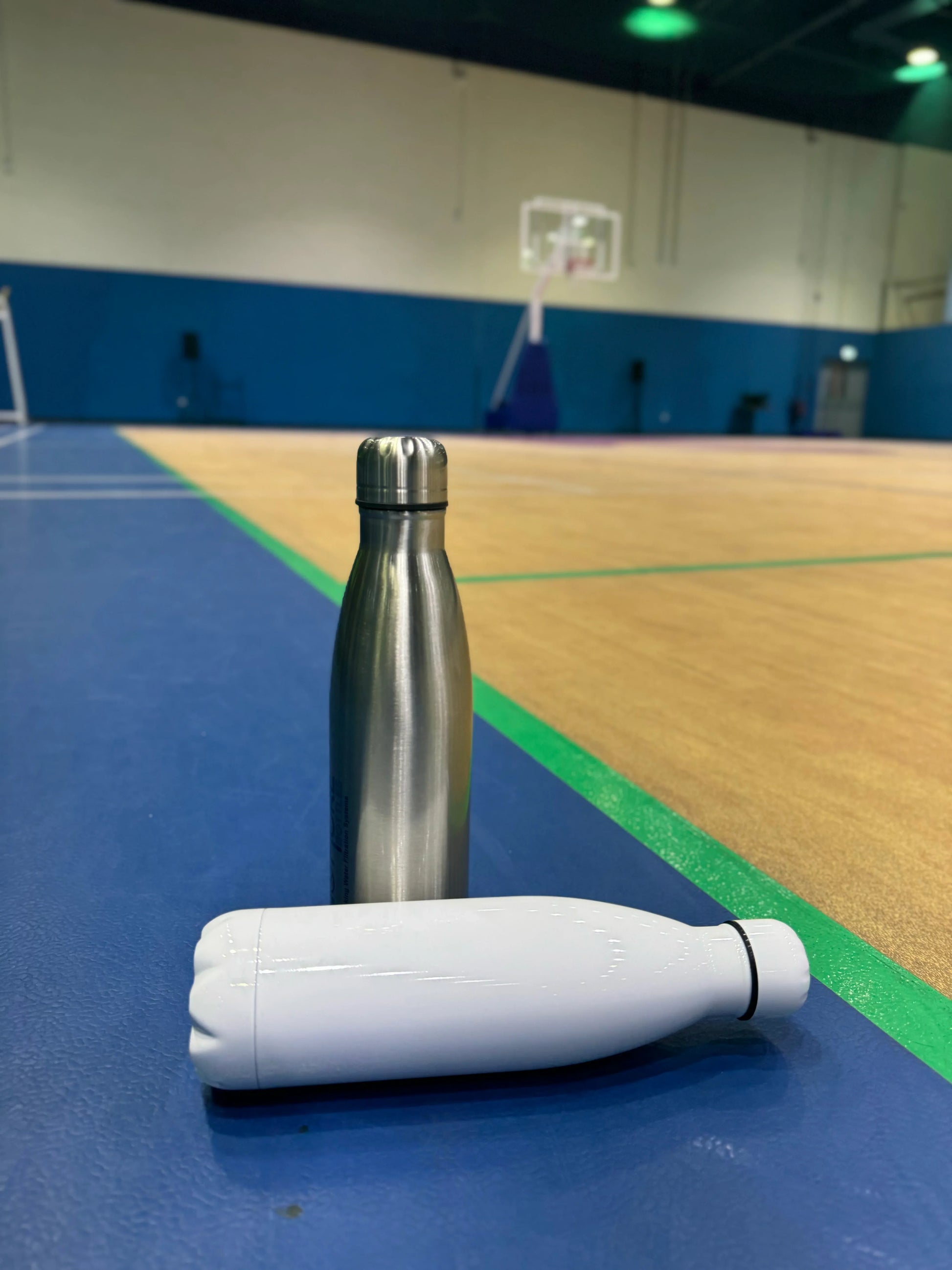 Two insulated bottles standing on the floor of an indoor basketball court, featuring Thermal Bottle 500ml.